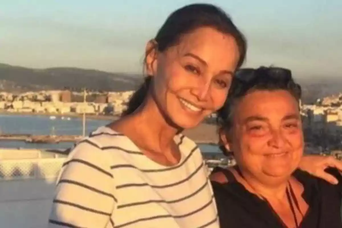 Dos mujeres sonríen abrazadas al aire libre con una ciudad y el mar de fondo Dos mujeres sonríen abrazadas al aire libre con una ciudad y el mar de fondo
