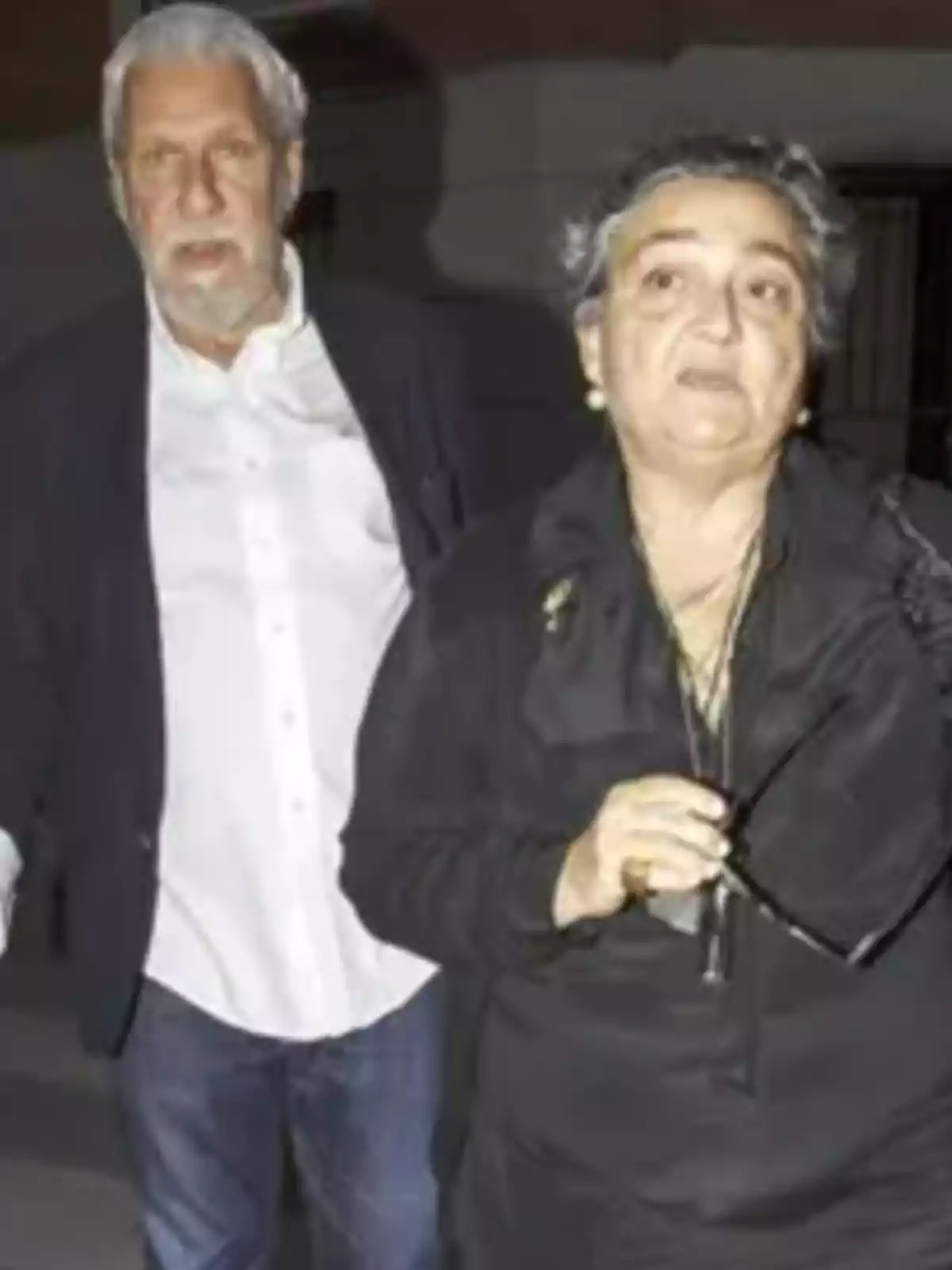 Una mujer de cabello corto y canoso vestida de negro sostiene unos lentes mientras un hombre de cabello blanco y barba la acompaña en el fondo Una mujer de cabello corto y canoso vestida de negro sostiene unos lentes mientras un hombre de cabello blanco y barba la acompaña en el fondo