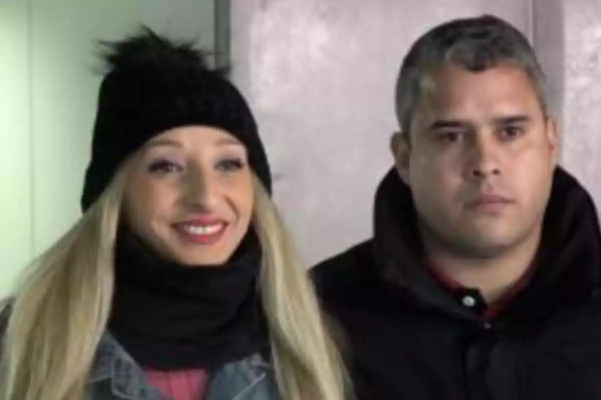 Una mujer rubia con gorro negro y bufanda sonríe junto a un hombre de cabello corto y expresión seria, ambos vestidos con ropa de invierno. Una mujer rubia con gorro negro y bufanda sonríe junto a un hombre de cabello corto y expresión seria, ambos vestidos con ropa de invierno.