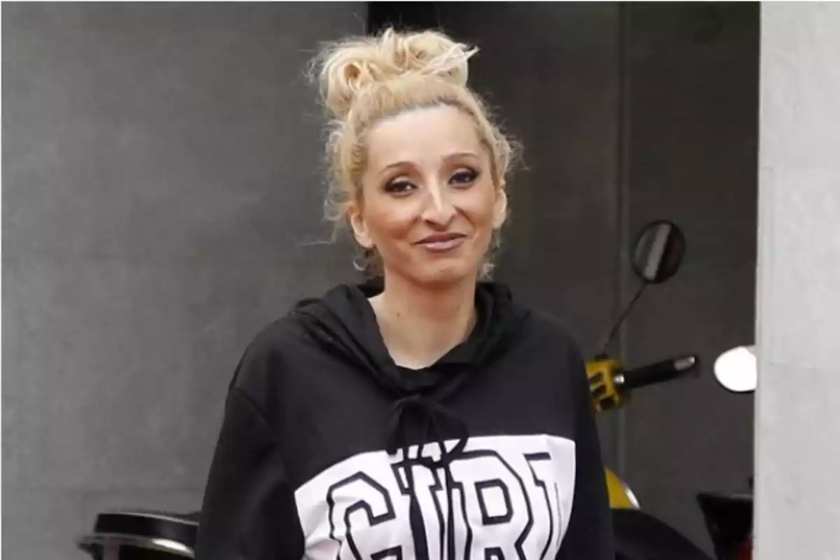 Mujer rubia con moño alto y sudadera negra con la palabra GIRL en letras grandes, sonriendo frente a una pared gris Mujer rubia con moño alto y sudadera negra con la palabra GIRL en letras grandes, sonriendo frente a una pared gris
