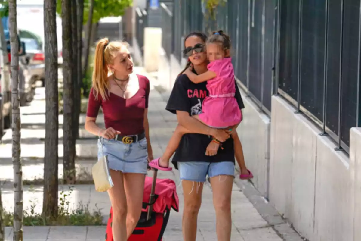 Dos mujeres caminan por la acera, una de ellas lleva una maleta roja y la otra carga a una niña vestida de rosa Dos mujeres caminan por la acera, una de ellas lleva una maleta roja y la otra carga a una niña vestida de rosa