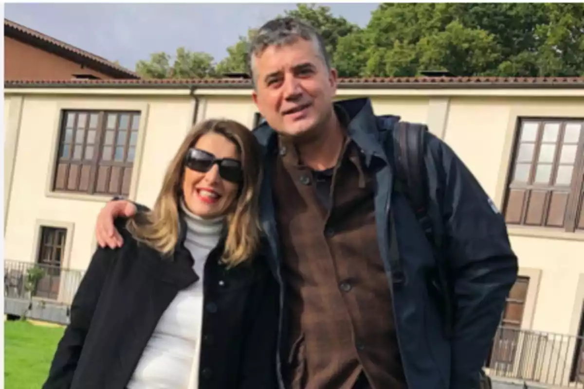 Una pareja sonriente posa al aire libre frente a un edificio de paredes claras y ventanas grandes rodeados de césped y árboles Una pareja sonriente posa al aire libre frente a un edificio de paredes claras y ventanas grandes rodeados de césped y árboles