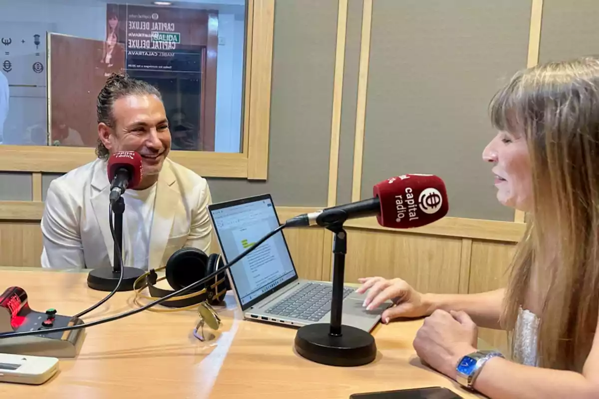 Dos personas conversan en una cabina de radio frente a micrófonos y una computadora portátil sobre la mesa Dos personas conversan en una cabina de radio frente a micrófonos y una computadora portátil sobre la mesa