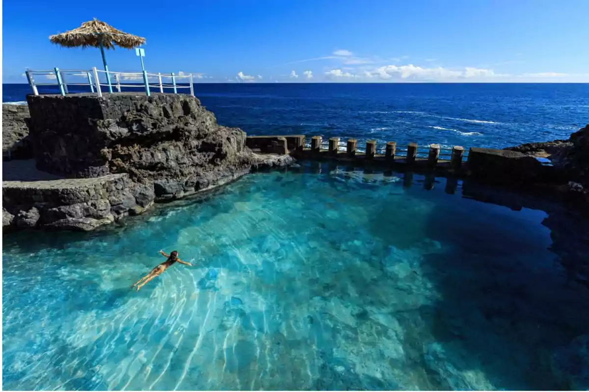 Mujer nadando en una piscina natural de agua cristalina junto al mar con un cielo despejado y una estructura de piedra con sombrilla de paja