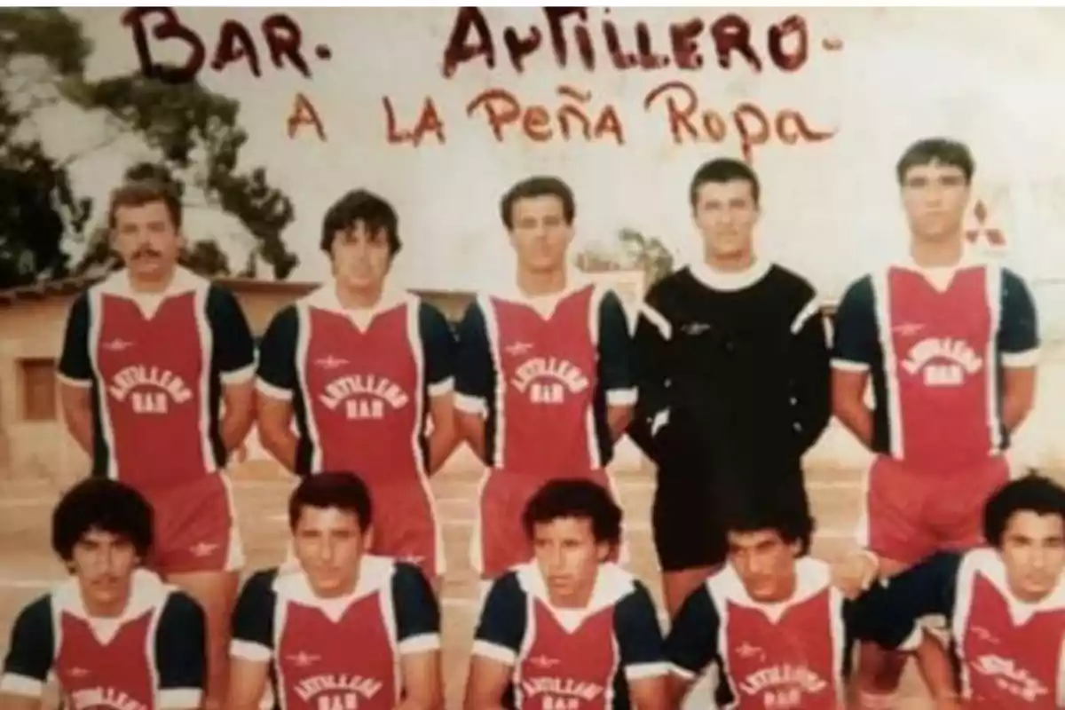 Equipo de fútbol posando para una foto grupal con uniformes rojos y azules que tienen la leyenda Artillero Bar, al fondo se observa una pared con la frase Bar Artillero a la Peña Ropa escrita a mano. Equipo de fútbol posando para una foto grupal con uniformes rojos y azules que tienen la leyenda Artillero Bar, al fondo se observa una pared con la frase Bar Artillero a la Peña Ropa escrita a mano.
