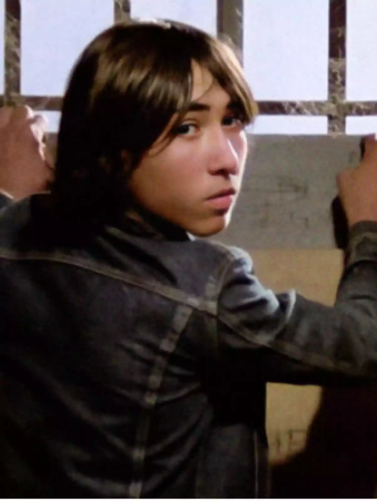 Joven de cabello castaño y chaqueta de mezclilla mirando hacia atrás mientras sostiene una reja Joven de cabello castaño y chaqueta de mezclilla mirando hacia atrás mientras sostiene una reja