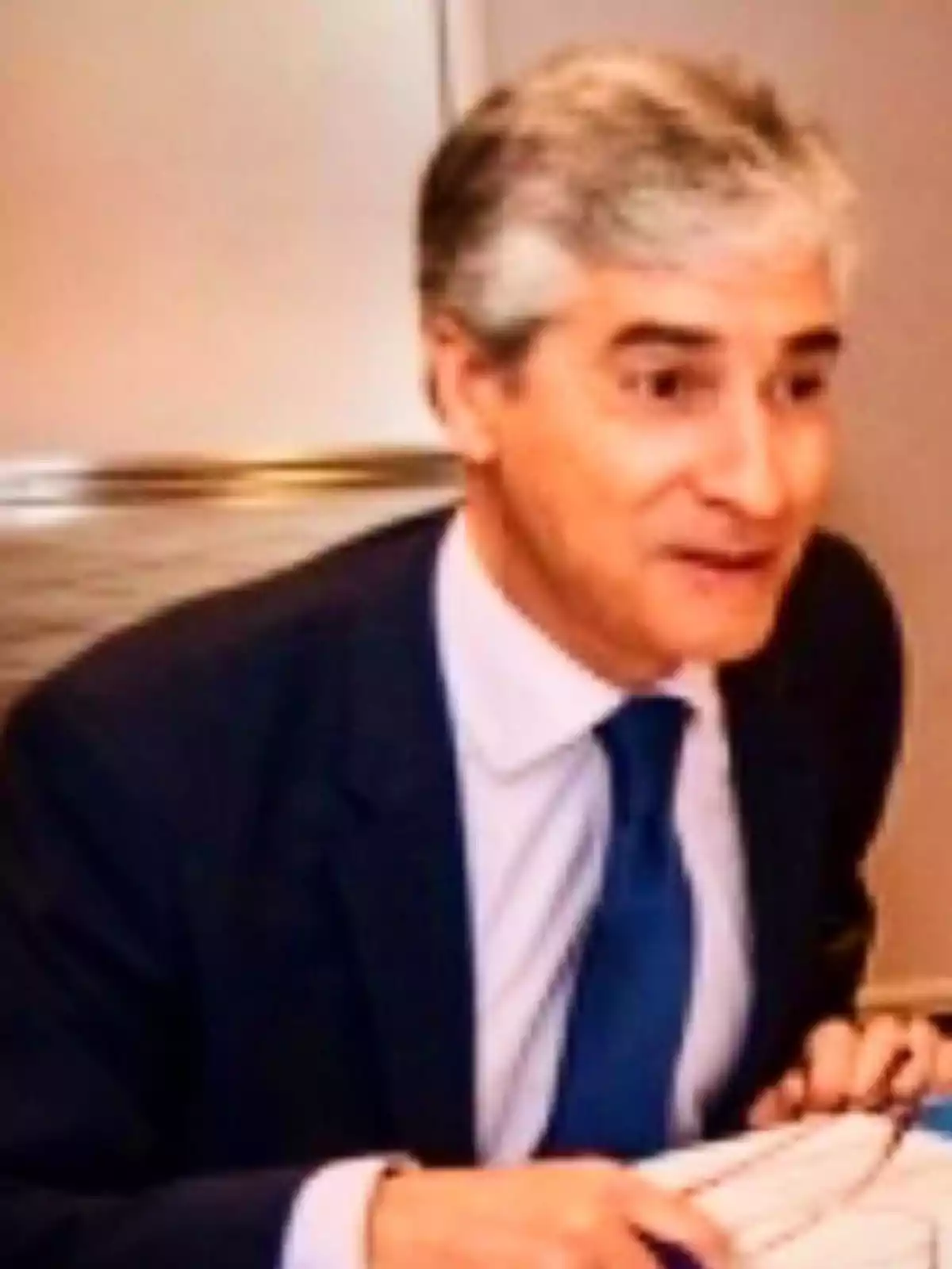 Hombre de cabello canoso con traje oscuro y corbata azul sentado en una mesa Hombre de cabello canoso con traje oscuro y corbata azul sentado en una mesa