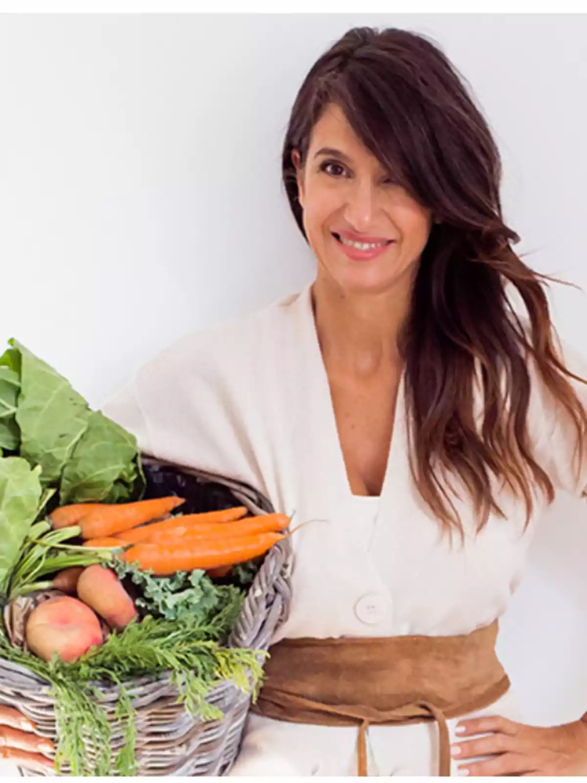 Mujer sonriente con cabello largo y oscuro sostiene una canasta de verduras frescas como zanahorias, papas y hojas verdes Mujer sonriente con cabello largo y oscuro sostiene una canasta de verduras frescas como zanahorias, papas y hojas verdes