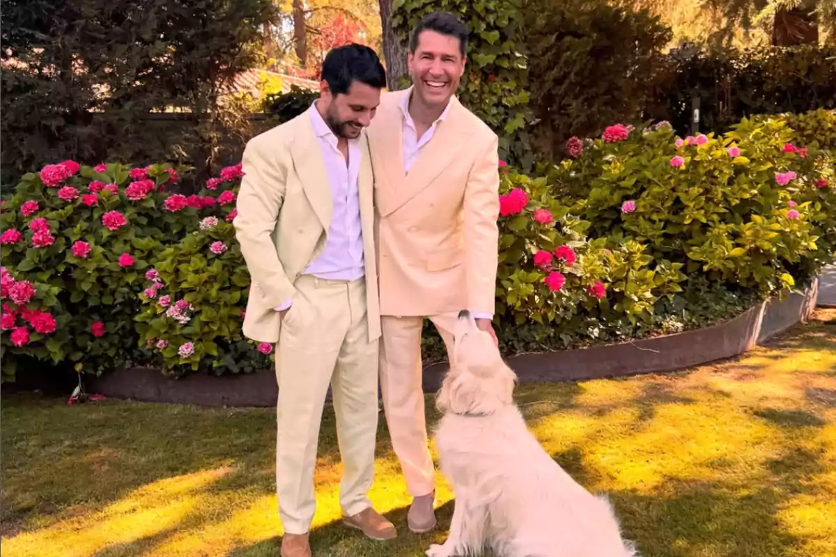Dos hombres vestidos con trajes claros sonríen en un jardín lleno de flores rosas mientras un perro blanco se sienta frente a ellos.