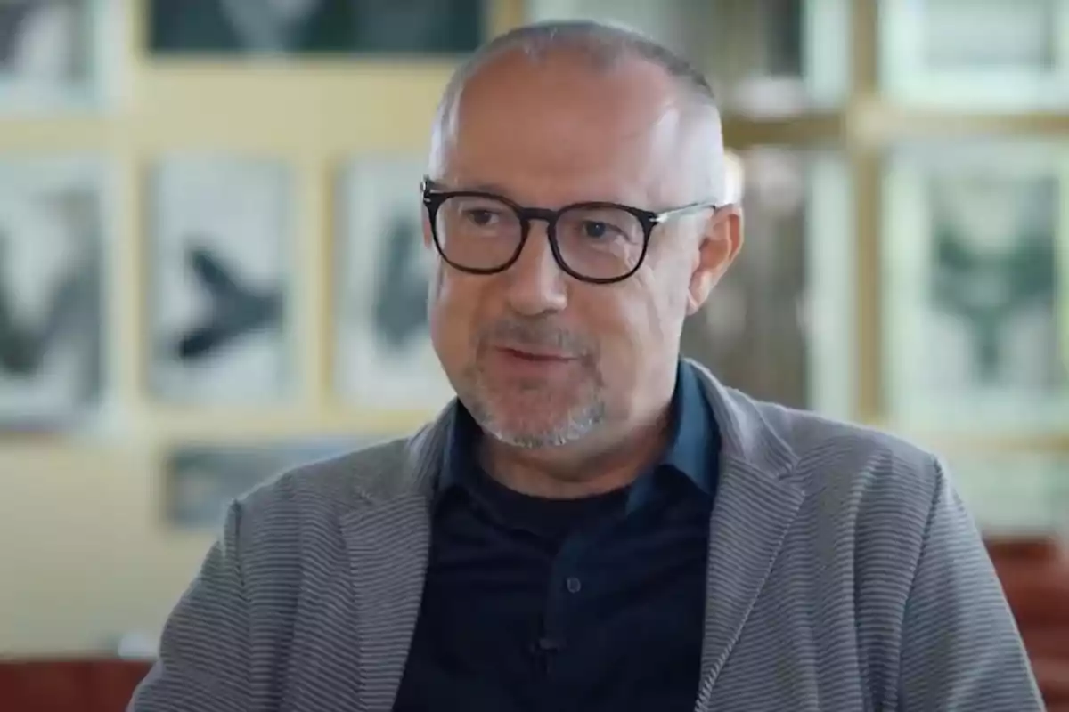 Hombre de mediana edad con gafas y barba corta, vestido con chaqueta gris y camisa azul, sentado en un ambiente interior con cuadros desenfocados al fondo Hombre de mediana edad con gafas y barba corta, vestido con chaqueta gris y camisa azul, sentado en un ambiente interior con cuadros desenfocados al fondo