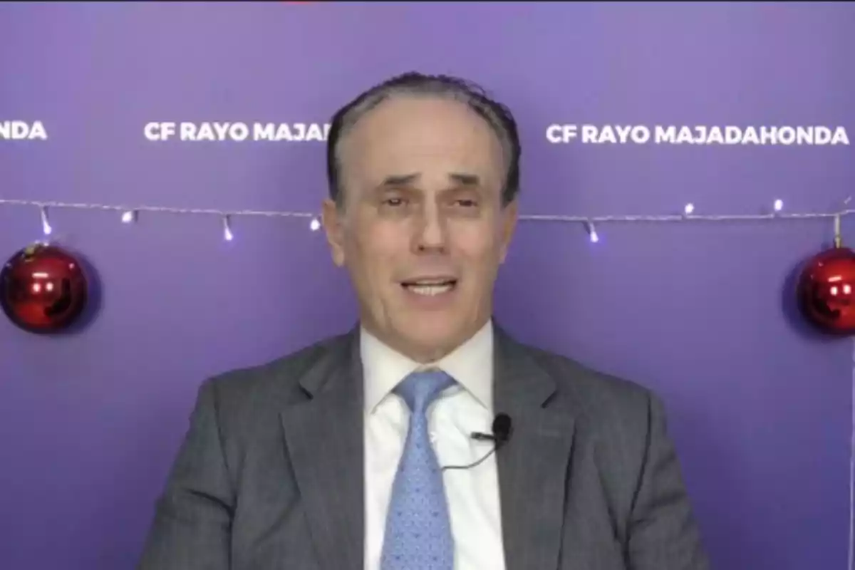 Hombre de traje y corbata hablando frente a un fondo morado decorado con luces y esferas rojas de Navidad y el texto CF Rayo Majadahonda Hombre de traje y corbata hablando frente a un fondo morado decorado con luces y esferas rojas de Navidad y el texto CF Rayo Majadahonda