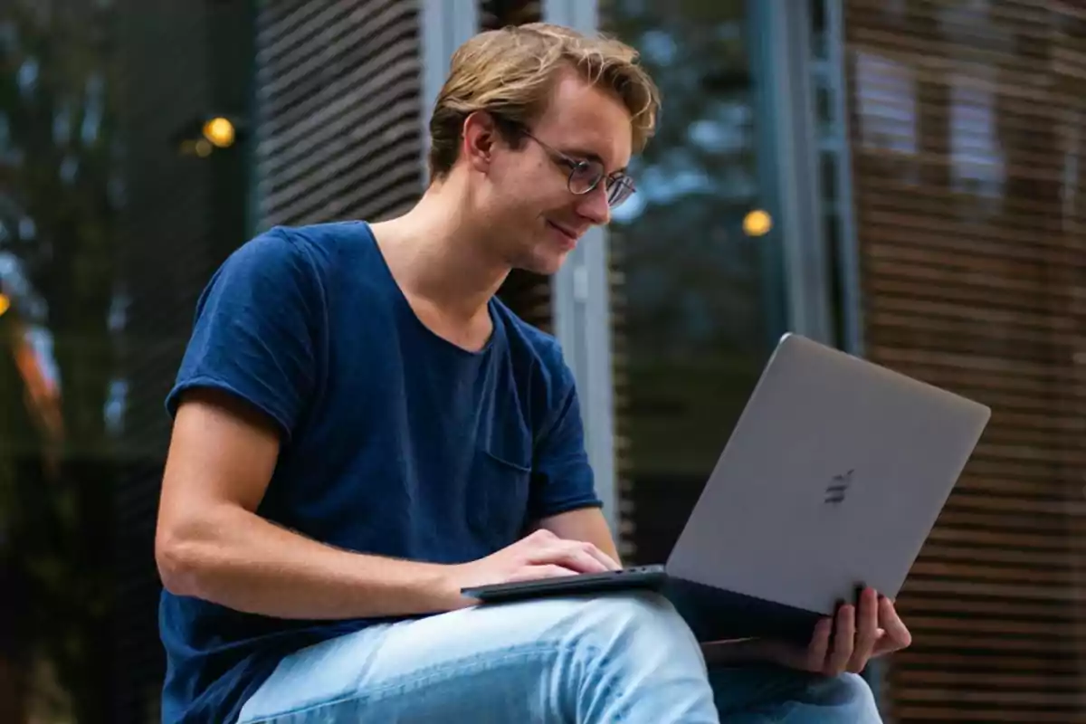 Joven con gafas usando una laptop plateada mientras está sentado al aire libre