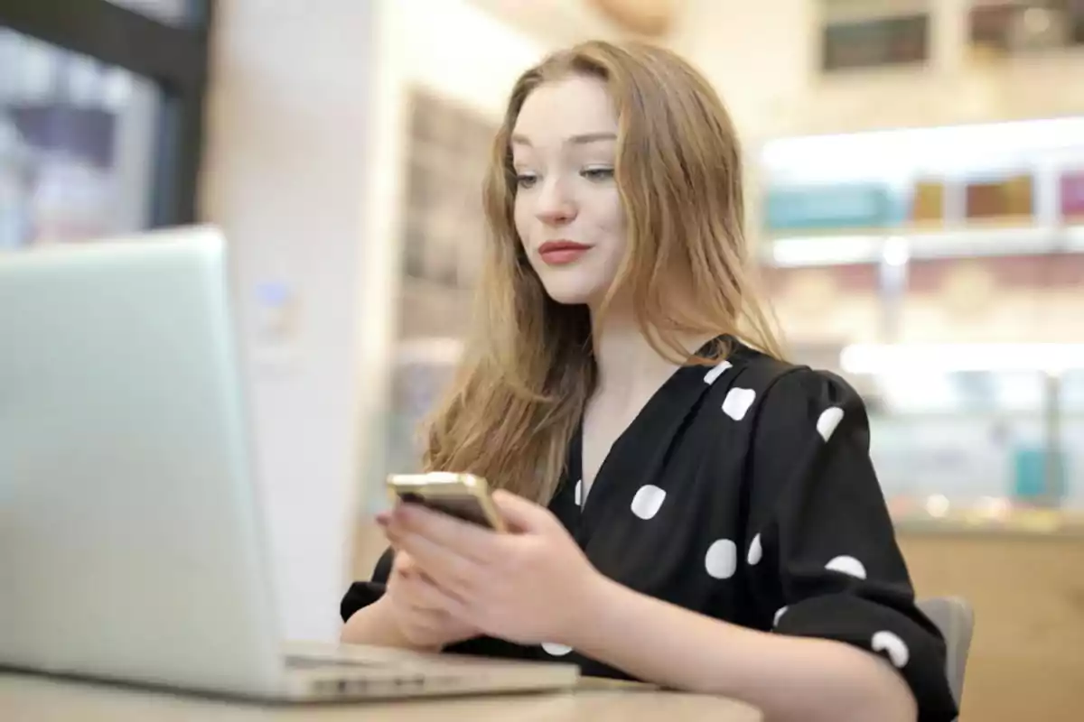 Mujer joven usando un teléfono móvil mientras trabaja en una computadora portátil en un ambiente interior iluminado Mujer joven usando un teléfono móvil mientras trabaja en una computadora portátil en un ambiente interior iluminado