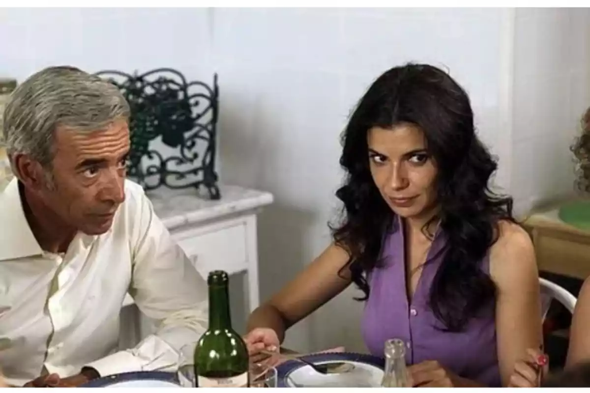 Dos personas sentadas a la mesa durante una comida, con expresiones serias y una botella de vino en primer plano Dos personas sentadas a la mesa durante una comida, con expresiones serias y una botella de vino en primer plano