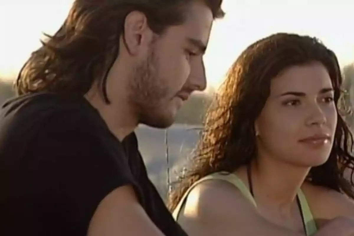Dos personas jóvenes sentadas al aire libre durante el atardecer, una mujer de cabello rizado y un hombre de cabello largo conversan en un ambiente tranquilo. Dos personas jóvenes sentadas al aire libre durante el atardecer, una mujer de cabello rizado y un hombre de cabello largo conversan en un ambiente tranquilo.