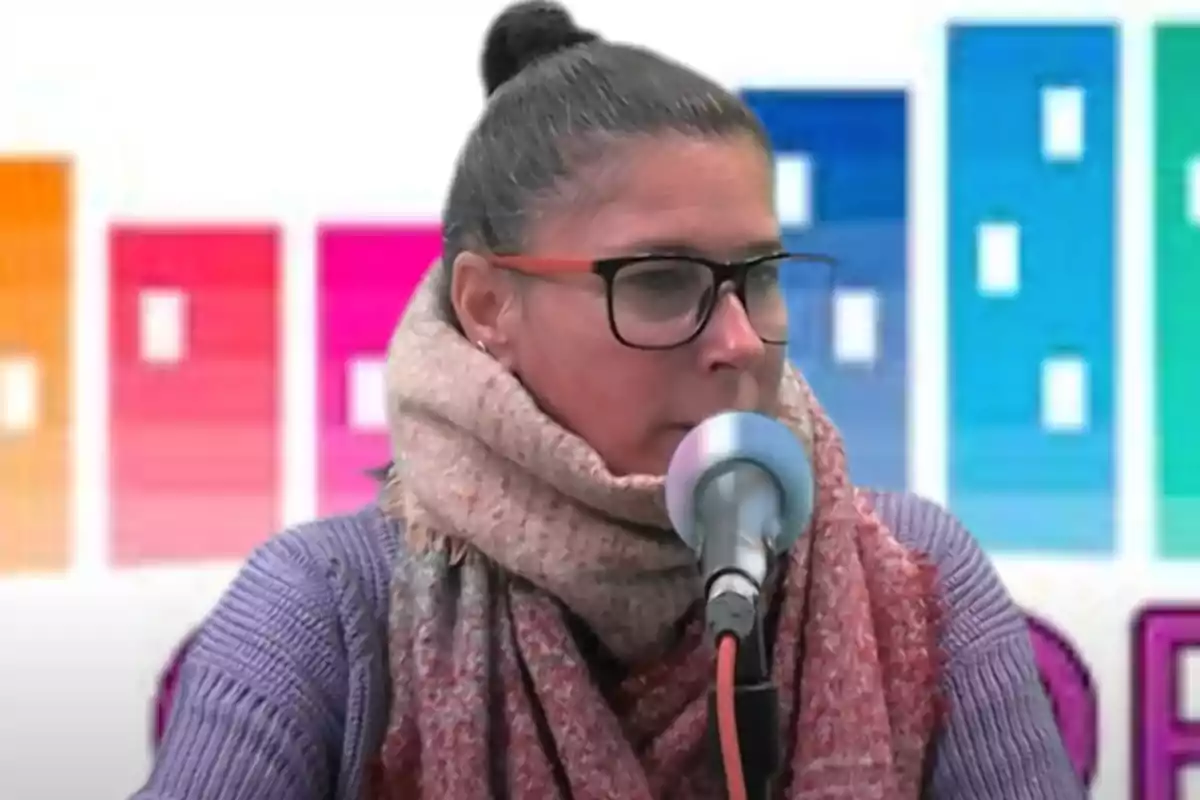 Mujer con gafas y bufanda hablando frente a un micrófono con fondo colorido de bloques rectangulares Mujer con gafas y bufanda hablando frente a un micrófono con fondo colorido de bloques rectangulares