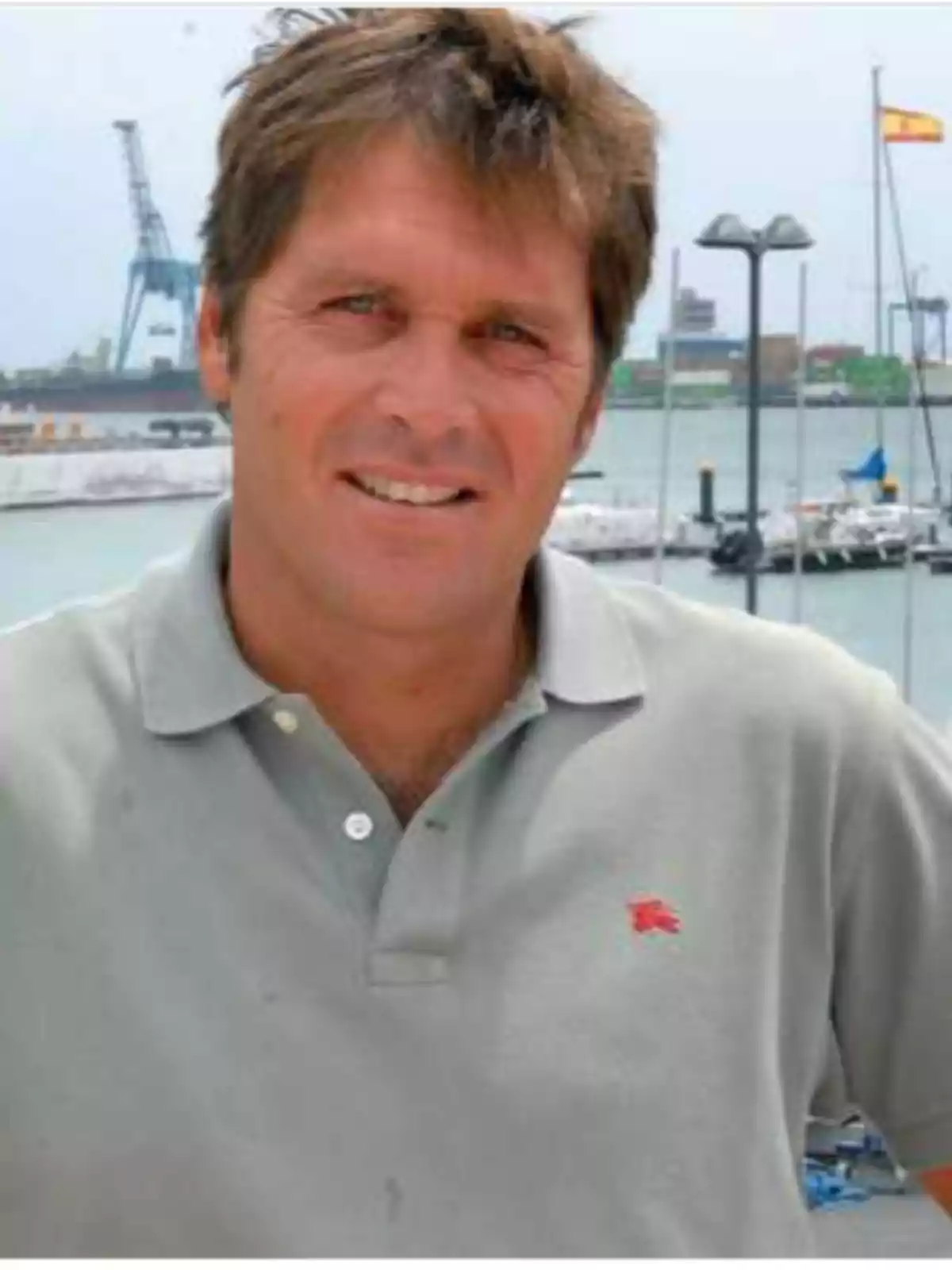 Brown-haired man wearing a gray t-jersey posing in front of a harbor with boats and a Spanish flag in the background