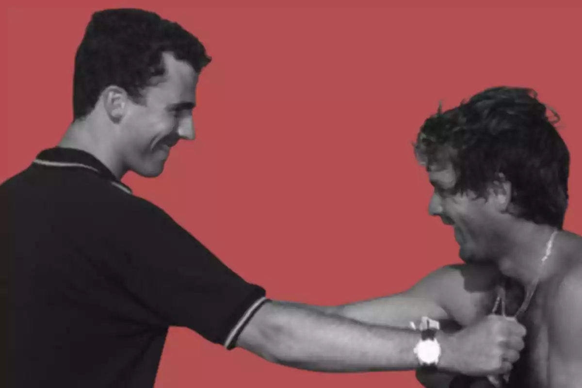 Two men smile as one of them holds the other’s chain against a red background.