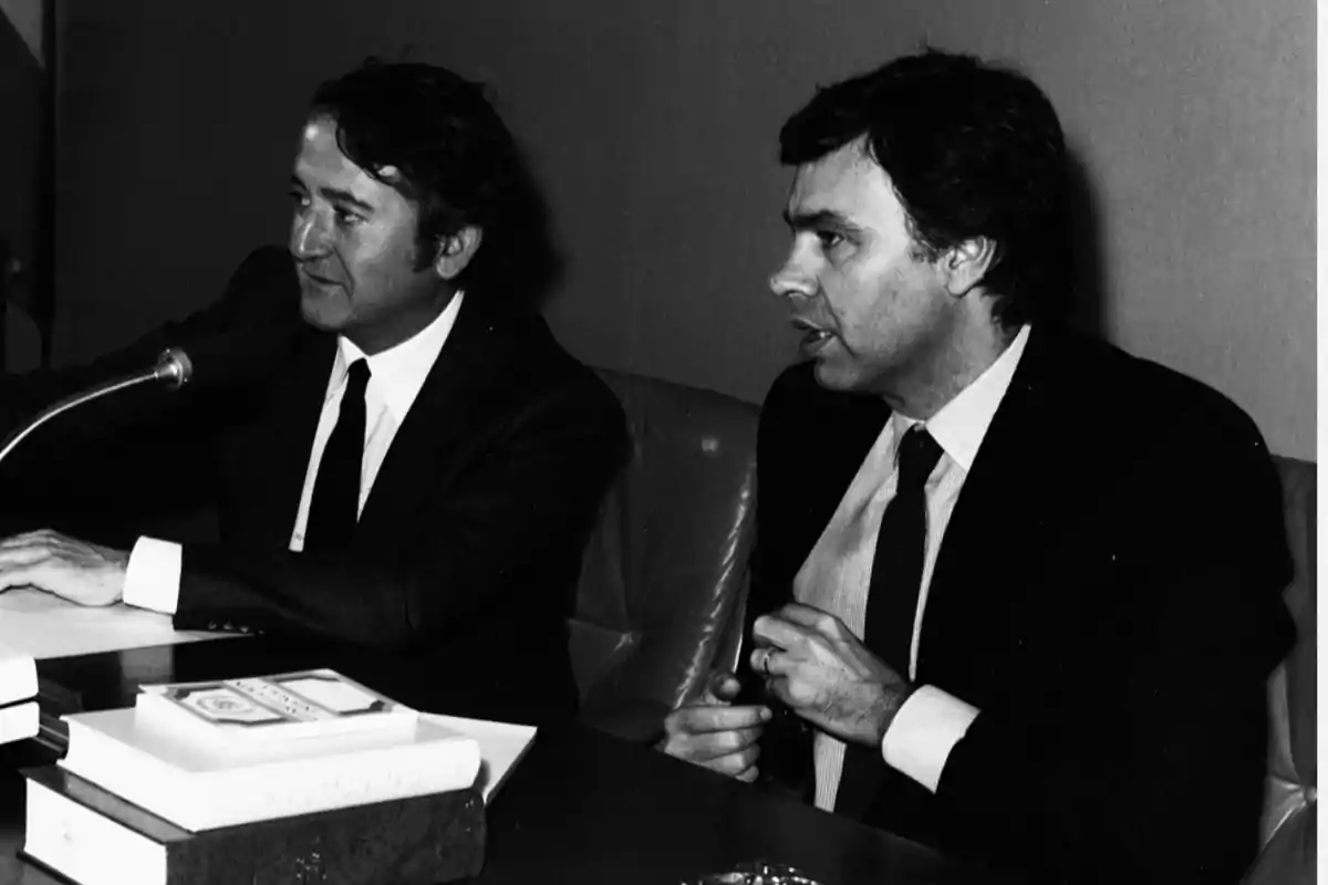 Dos hombres con traje y corbata sentados en una mesa durante una reunión o conferencia, uno de ellos hablando frente a un micrófono y varios libros apilados en primer plano Dos hombres con traje y corbata sentados en una mesa durante una reunión o conferencia, uno de ellos hablando frente a un micrófono y varios libros apilados en primer plano