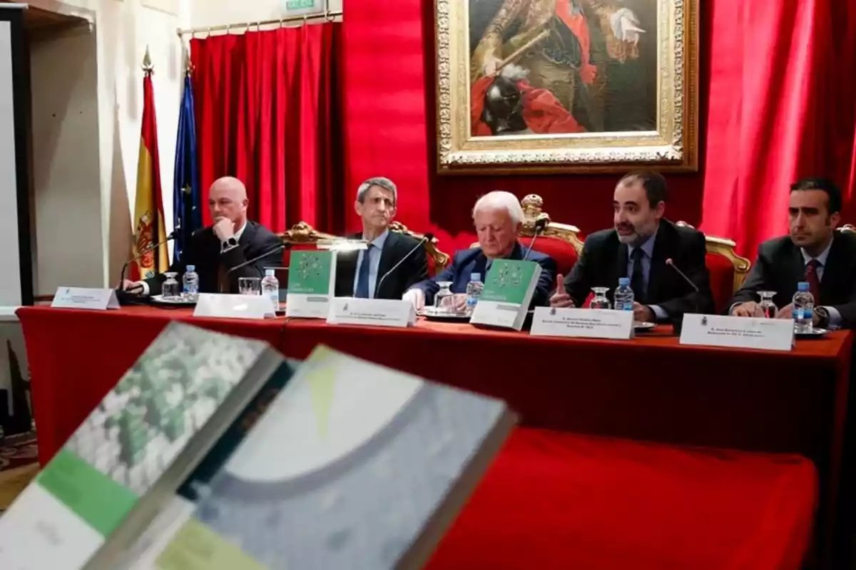 Cinco hombres sentados en una mesa larga con manteles rojos durante una conferencia o presentación de libros, con banderas y un cuadro en el fondo Cinco hombres sentados en una mesa larga con manteles rojos durante una conferencia o presentación de libros, con banderas y un cuadro en el fondo