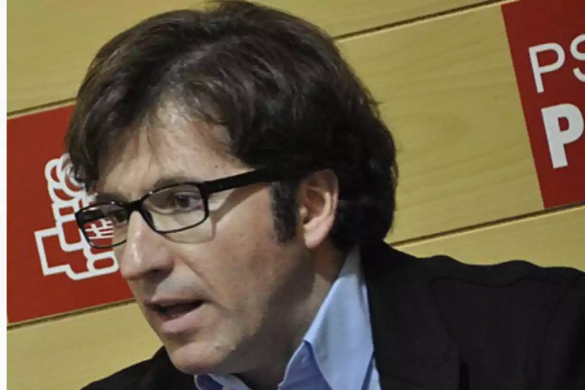 Hombre con gafas y cabello castaño hablando frente a un fondo con logotipos y letras rojas Hombre con gafas y cabello castaño hablando frente a un fondo con logotipos y letras rojas