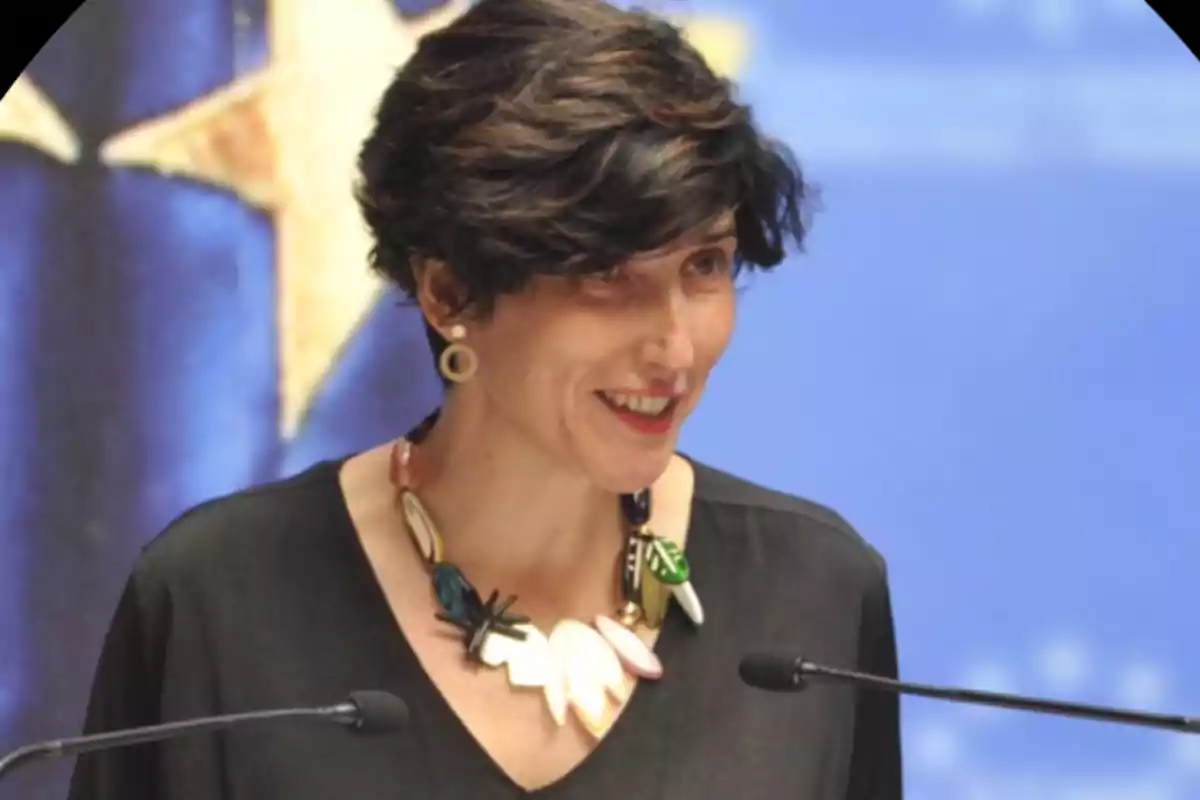 Mujer de cabello corto y oscuro hablando frente a dos micrófonos con un fondo azul y una estrella amarilla desenfocada Mujer de cabello corto y oscuro hablando frente a dos micrófonos con un fondo azul y una estrella amarilla desenfocada