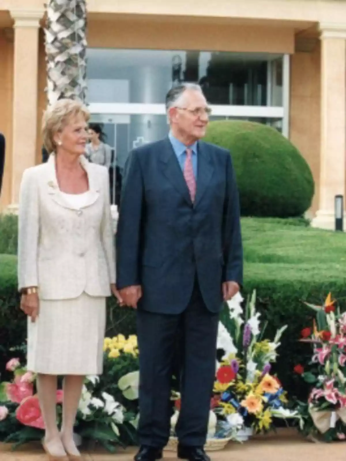 Una pareja de personas mayores elegantemente vestidas está de pie al aire libre, rodeada de arreglos florales y arbustos recortados, frente a un edificio. Una pareja de personas mayores elegantemente vestidas está de pie al aire libre, rodeada de arreglos florales y arbustos recortados, frente a un edificio.