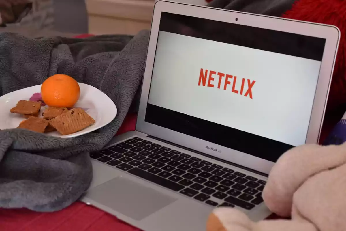 Una laptop con la pantalla mostrando el logo de Netflix, junto a un plato con galletas y una naranja sobre una manta gris. Una laptop con la pantalla mostrando el logo de Netflix, junto a un plato con galletas y una naranja sobre una manta gris.
