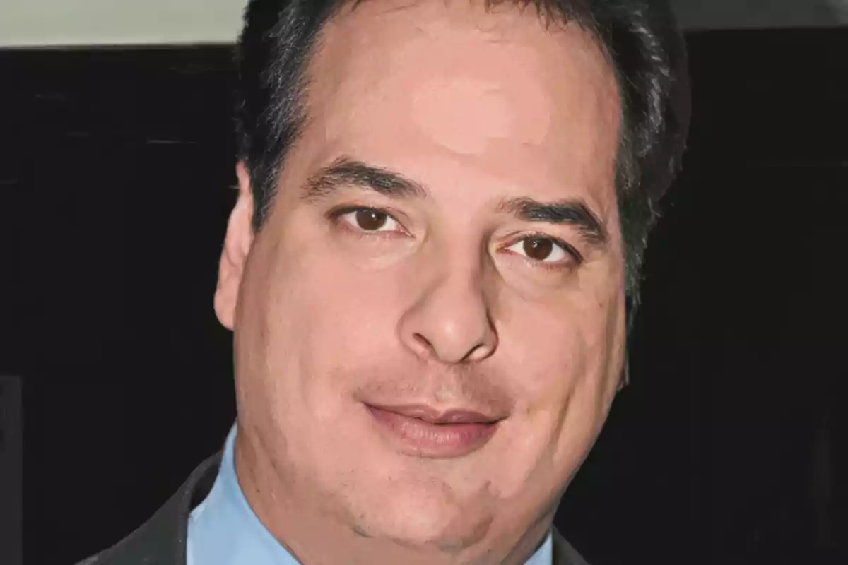 Hombre de cabello oscuro y piel clara con expresión seria, usando camisa azul y saco oscuro, fondo negro. Hombre de cabello oscuro y piel clara con expresión seria, usando camisa azul y saco oscuro, fondo negro.