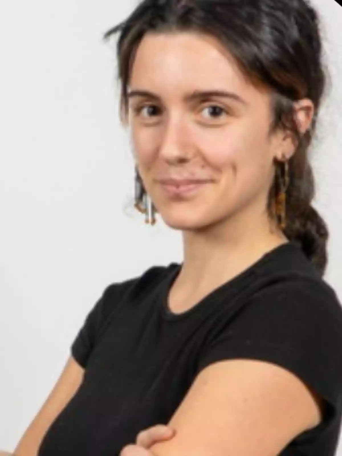 Mujer joven de cabello oscuro recogido, con camiseta negra y pendientes largos, mirando a la cámara y sonriendo levemente Mujer joven de cabello oscuro recogido, con camiseta negra y pendientes largos, mirando a la cámara y sonriendo levemente