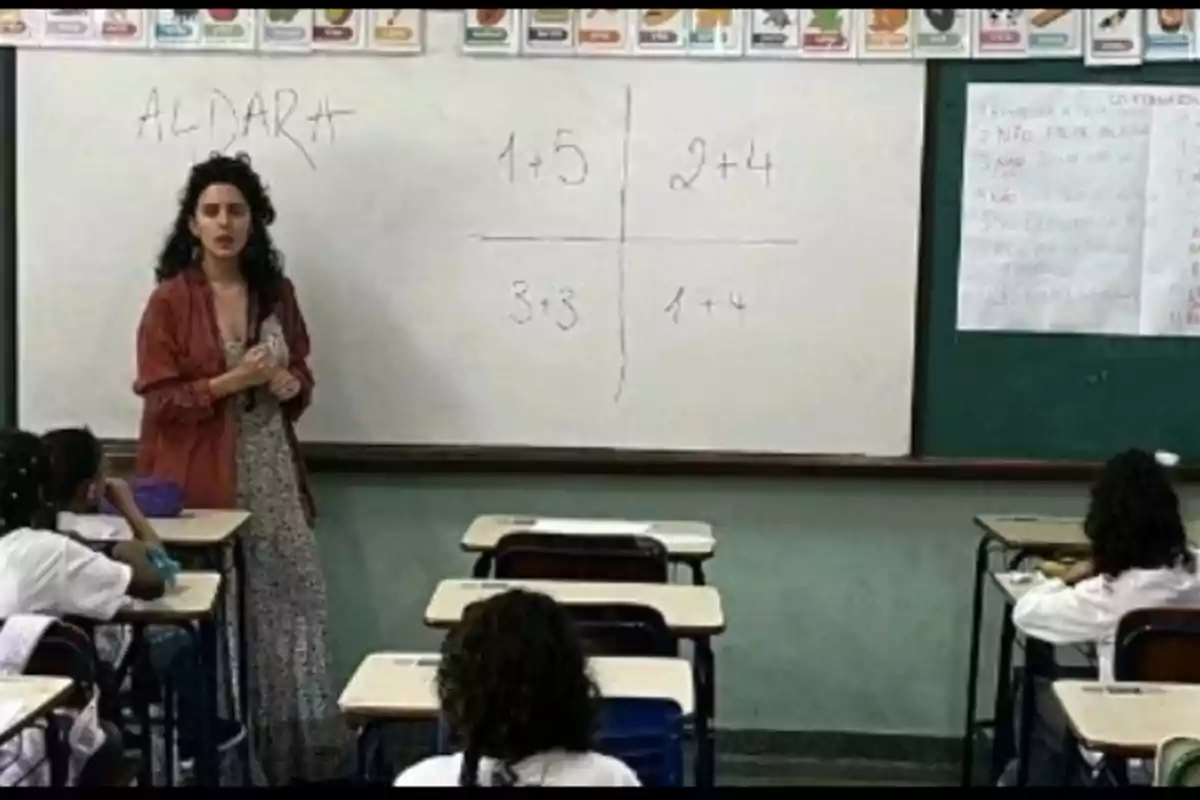 Una maestra está de pie junto a un pizarrón en un salón de clases mientras varios niños sentados en sus pupitres observan y escuchan la lección de matemáticas escrita en el pizarrón Una maestra está de pie junto a un pizarrón en un salón de clases mientras varios niños sentados en sus pupitres observan y escuchan la lección de matemáticas escrita en el pizarrón