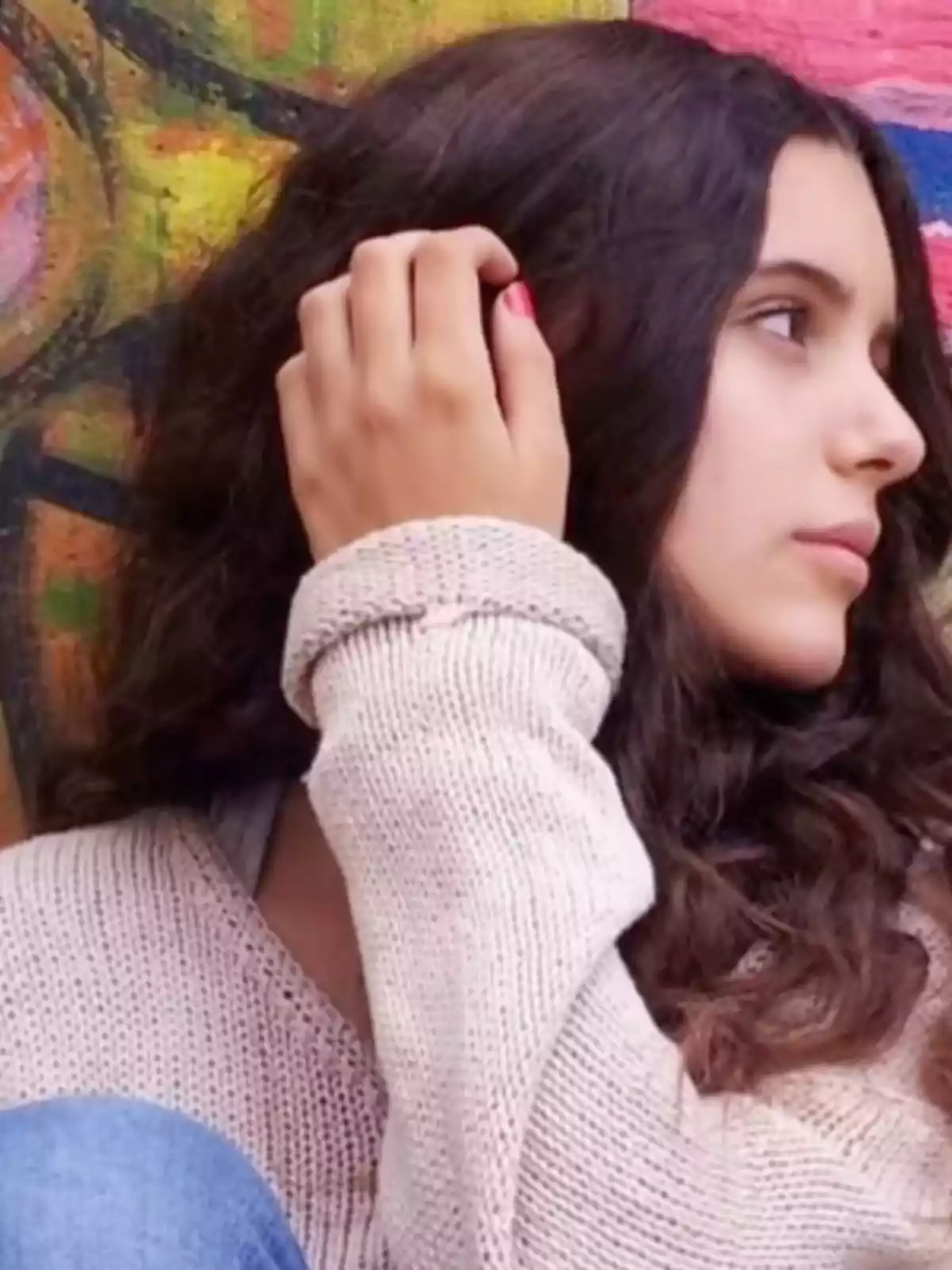 Joven de cabello largo y ondulado con suéter beige sentada frente a una pared colorida mirando hacia la derecha Joven de cabello largo y ondulado con suéter beige sentada frente a una pared colorida mirando hacia la derecha