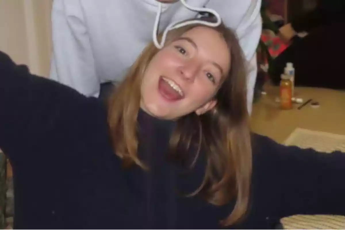 Joven sonriente con los brazos extendidos y un suéter oscuro, con una persona detrás sosteniendo una cuerda blanca sobre su cabeza en un ambiente interior. Joven sonriente con los brazos extendidos y un suéter oscuro, con una persona detrás sosteniendo una cuerda blanca sobre su cabeza en un ambiente interior.