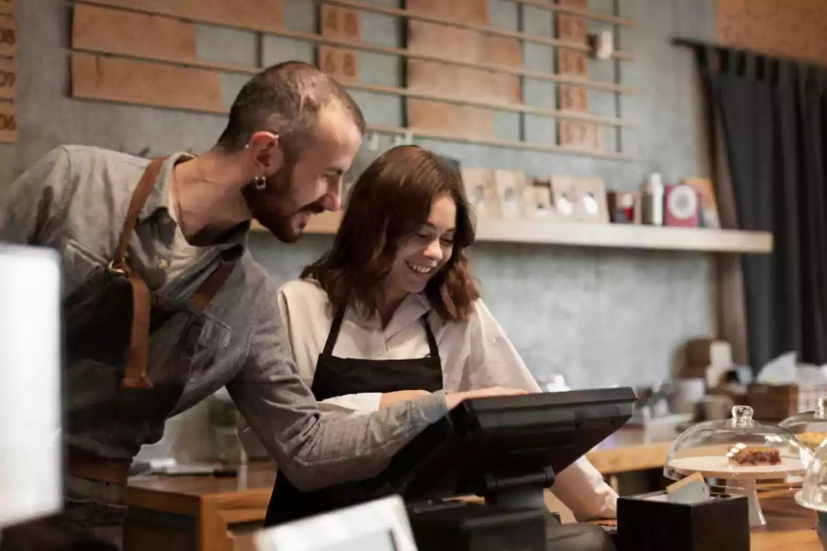 Dos personas con delantales sonríen mientras usan una caja registradora en una cafetería