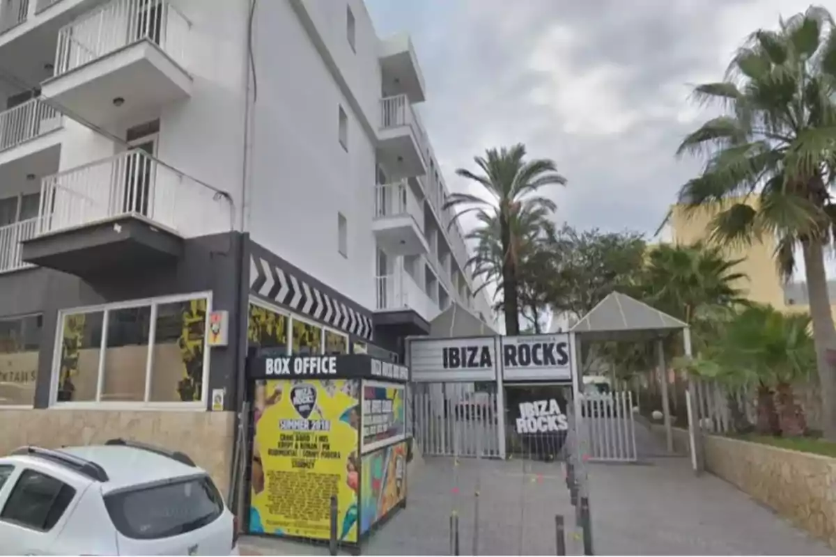 Entrada principal del hotel Ibiza Rocks con palmeras, un coche blanco estacionado y carteles promocionales en la entrada.