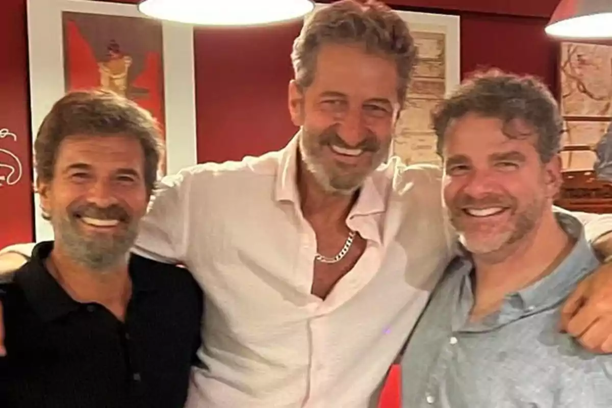 Tres hombres sonrientes posan juntos en un ambiente interior con paredes rojas y cuadros en el fondo Tres hombres sonrientes posan juntos en un ambiente interior con paredes rojas y cuadros en el fondo