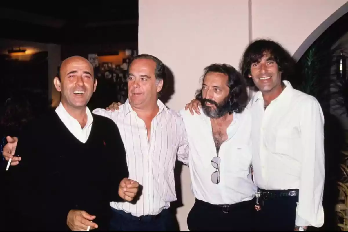 Cuatro hombres posan juntos y sonríen en un ambiente interior, dos de ellos sostienen cigarrillos y todos visten ropa casual clara Cuatro hombres posan juntos y sonríen en un ambiente interior, dos de ellos sostienen cigarrillos y todos visten ropa casual clara