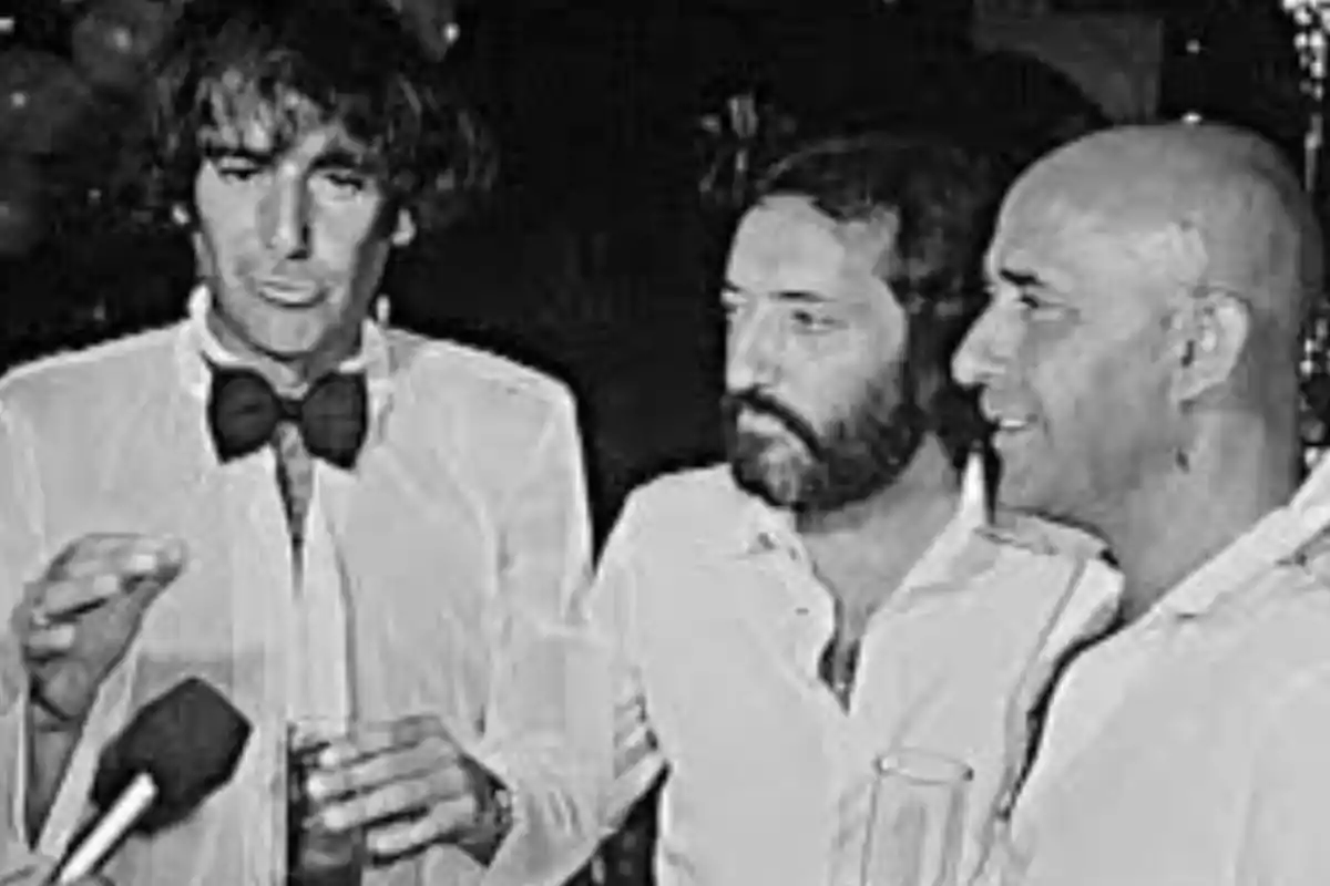 Tres hombres vestidos de manera elegante conversan y sostienen bebidas en un ambiente social Tres hombres vestidos de manera elegante conversan y sostienen bebidas en un ambiente social