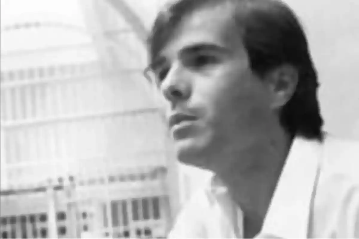 Hombre joven de cabello oscuro y corto con camisa clara en una imagen en blanco y negro, fondo desenfocado con estructuras metálicas. Hombre joven de cabello oscuro y corto con camisa clara en una imagen en blanco y negro, fondo desenfocado con estructuras metálicas.