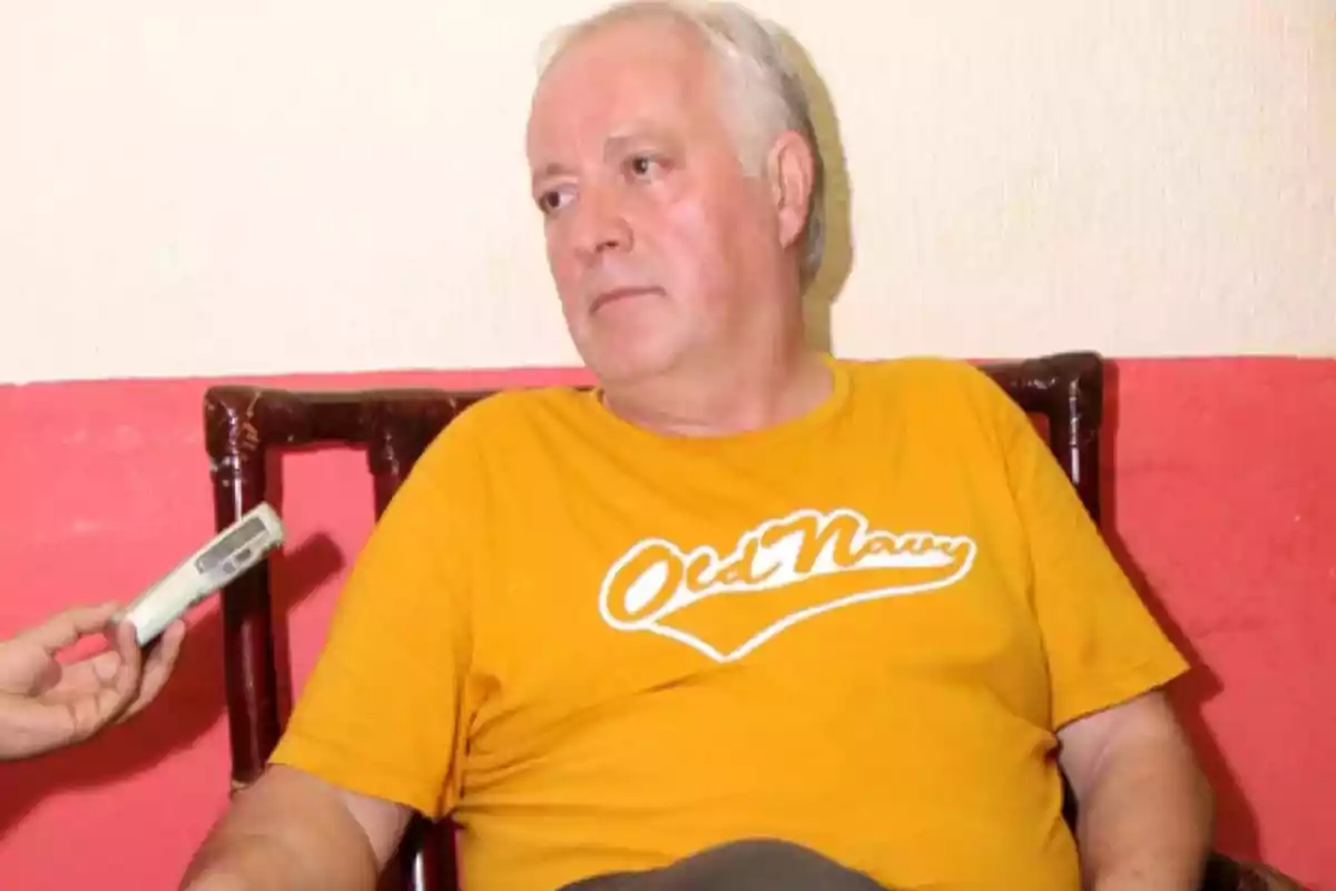 Hombre de cabello canoso con camiseta amarilla sentado en una silla mientras le hacen una entrevista con una grabadora Hombre de cabello canoso con camiseta amarilla sentado en una silla mientras le hacen una entrevista con una grabadora