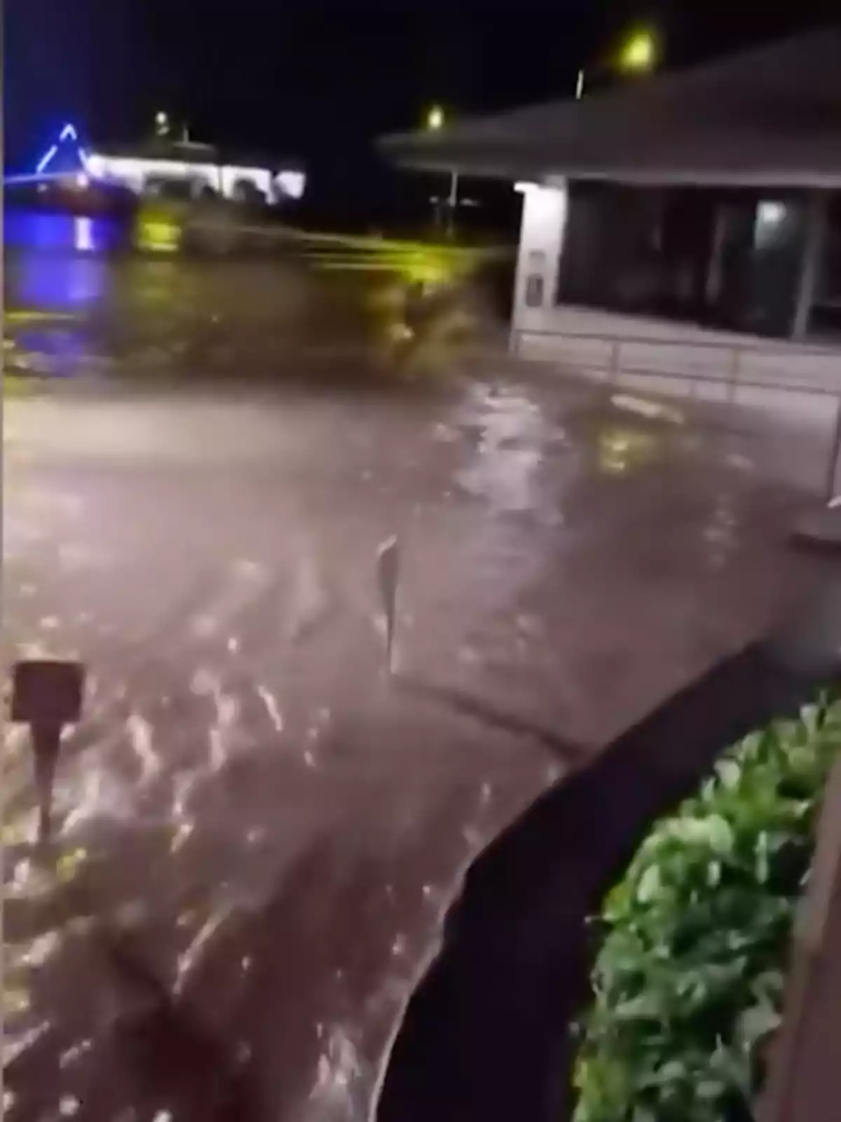 Calle inundada por una fuerte corriente de agua durante la noche con luces reflejadas en el agua y un edificio al fondo Calle inundada por una fuerte corriente de agua durante la noche con luces reflejadas en el agua y un edificio al fondo