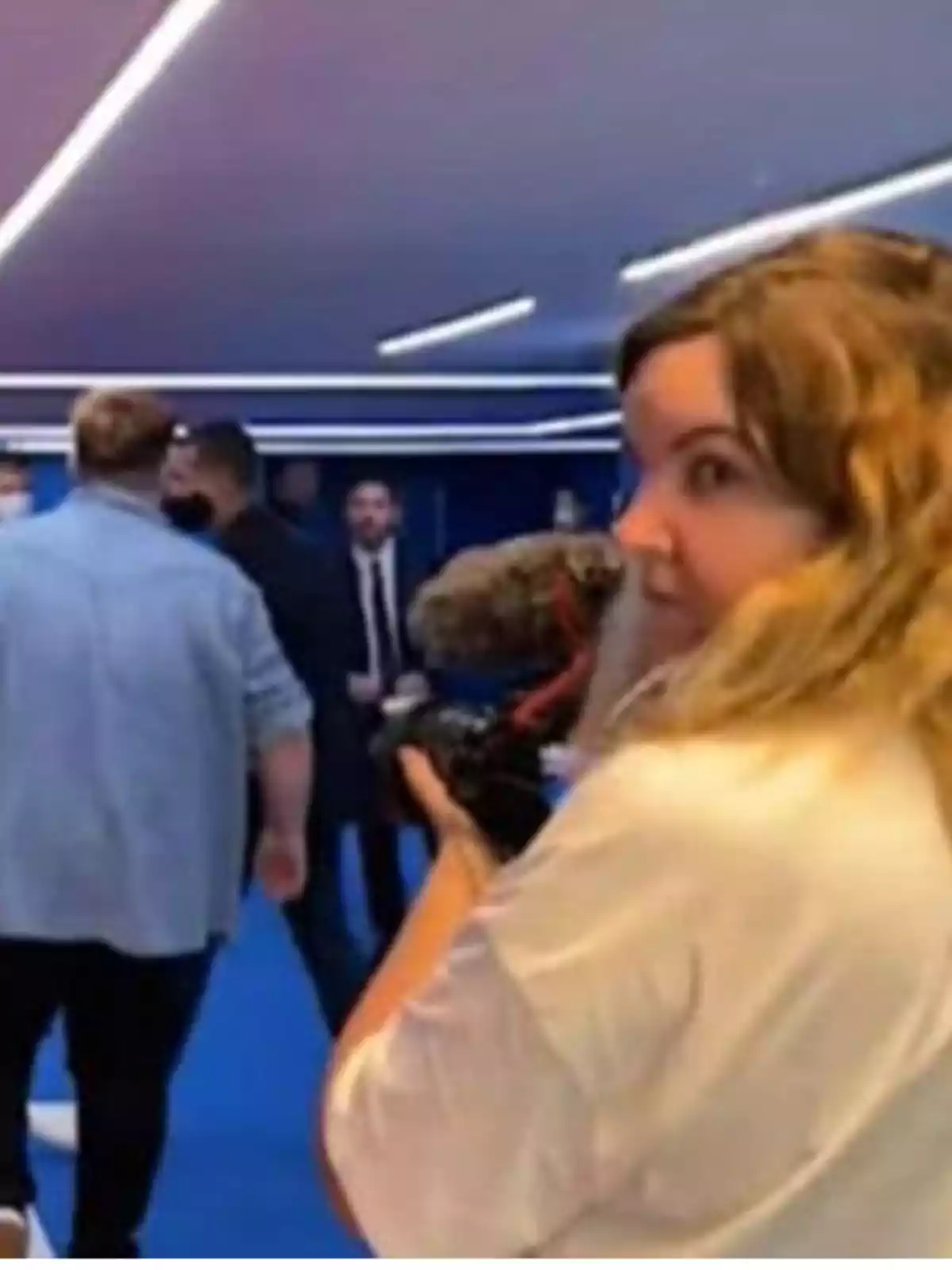 Mujer con cabello rubio sosteniendo una cámara en un evento con varias personas y luces en el techo Mujer con cabello rubio sosteniendo una cámara en un evento con varias personas y luces en el techo