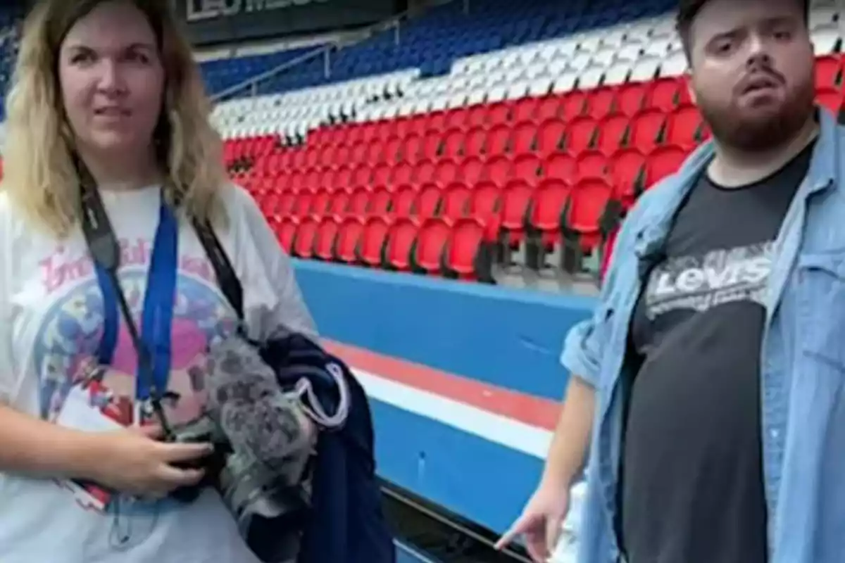 Dos personas de pie en un estadio con asientos rojos, blancos y azules, una de ellas sostiene una cámara y la otra viste una camiseta negra con chaqueta de mezclilla. Dos personas de pie en un estadio con asientos rojos, blancos y azules, una de ellas sostiene una cámara y la otra viste una camiseta negra con chaqueta de mezclilla.