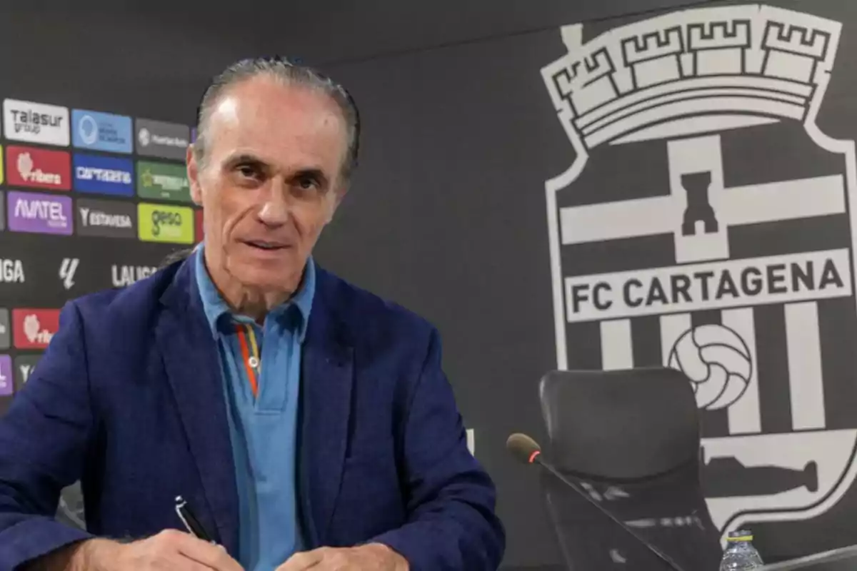 Hombre de cabello canoso firmando un documento en una sala de prensa con el escudo de FC Cartagena al fondo