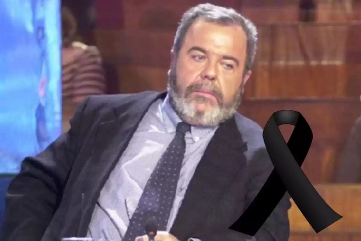 Hombre de barba canosa con traje y corbata sentado en un estudio de televisión con un moño negro de luto en la imagen.