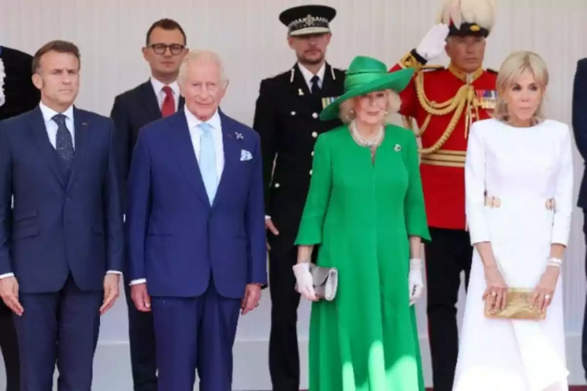 Four elegantly dressed people pose for an official photo alongside security staff members and a guard in a red uniform. Four elegantly dressed people pose for an official photo alongside security staff members and a guard in a red uniform.