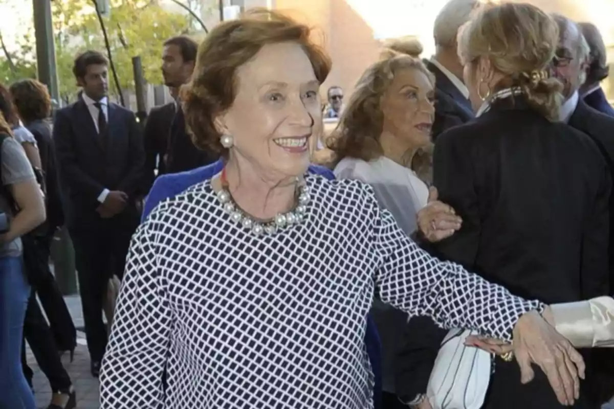 Mujer mayor sonriente con collar de perlas en un evento social al aire libre rodeada de otras personas Mujer mayor sonriente con collar de perlas en un evento social al aire libre rodeada de otras personas
