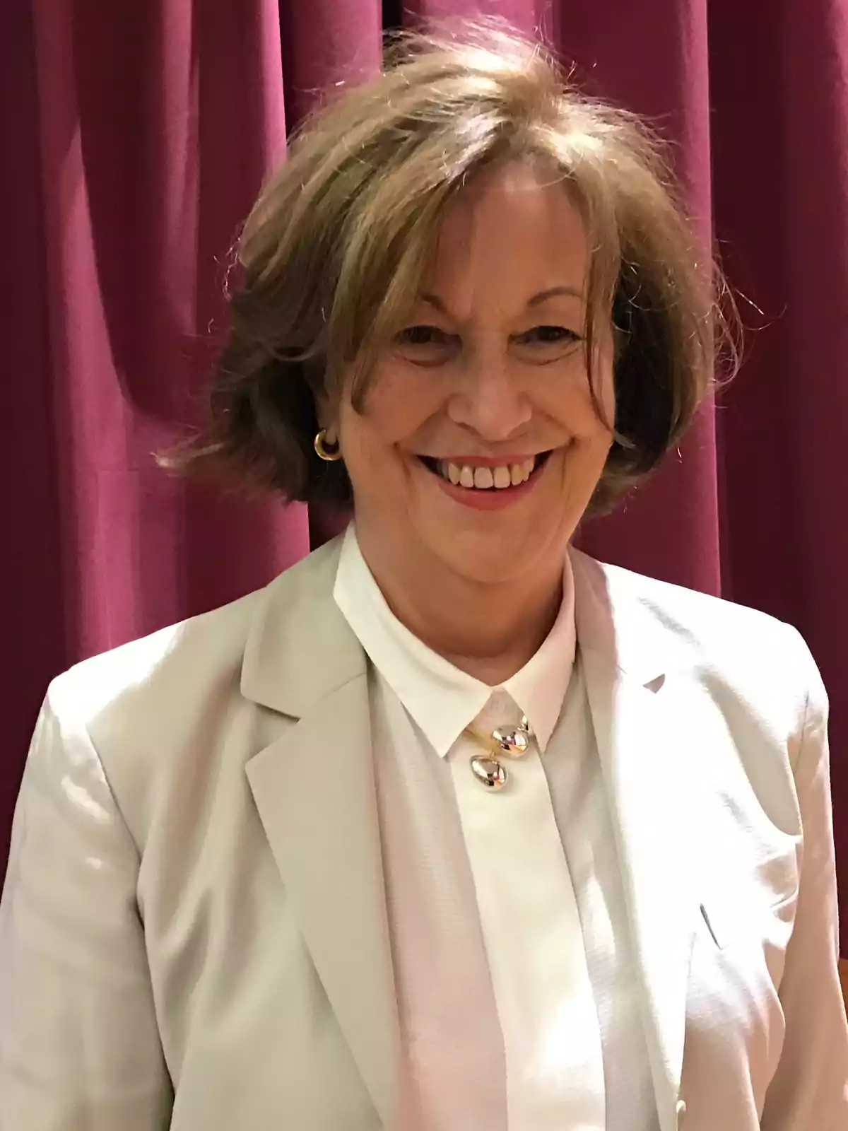 Mujer de cabello corto y castaño sonriendo, vestida con saco y blusa blanca, posando frente a una cortina color vino Mujer de cabello corto y castaño sonriendo, vestida con saco y blusa blanca, posando frente a una cortina color vino