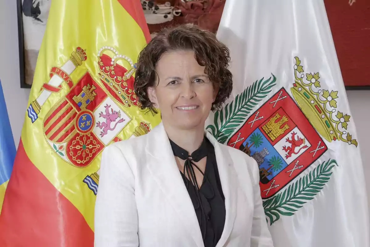 Mujer de cabello corto y rizado con saco blanco posando frente a las banderas de España y de una comunidad autónoma Mujer de cabello corto y rizado con saco blanco posando frente a las banderas de España y de una comunidad autónoma