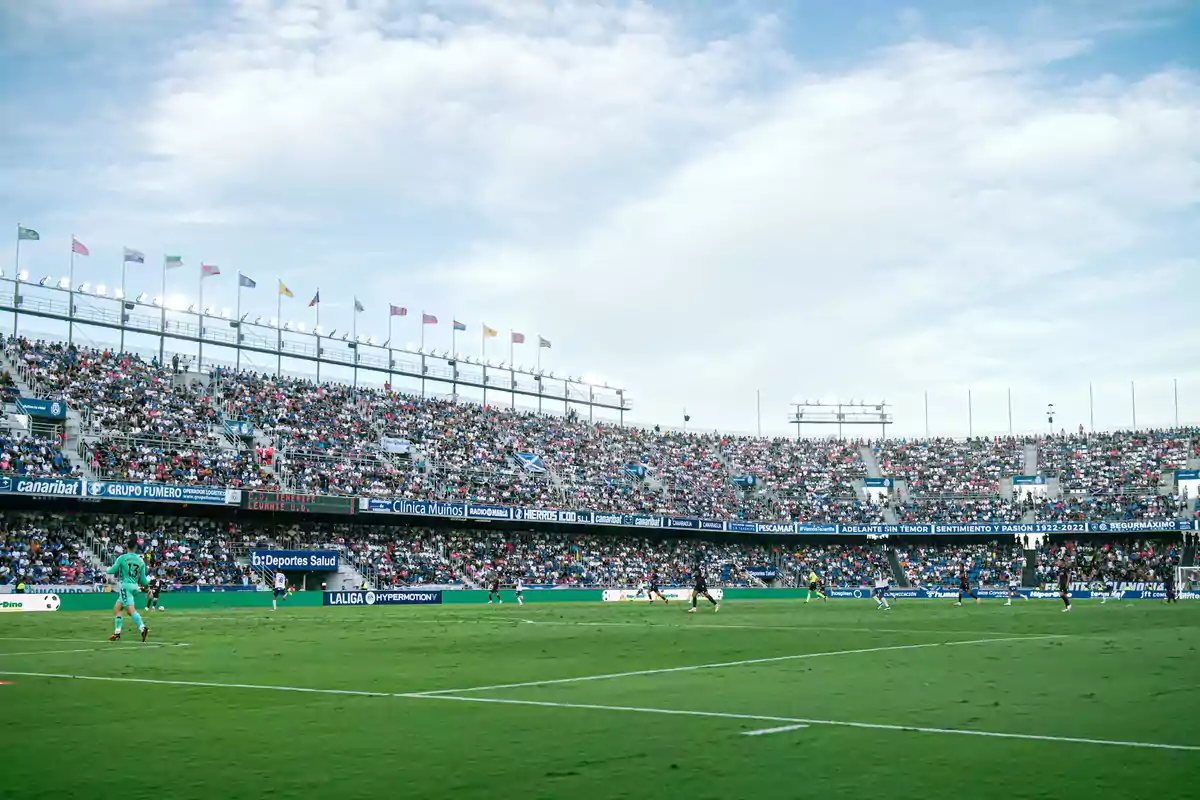 Estadio de fútbol lleno de aficionados durante un partido bajo un cielo parcialmente nublado Estadio de fútbol lleno de aficionados durante un partido bajo un cielo parcialmente nublado